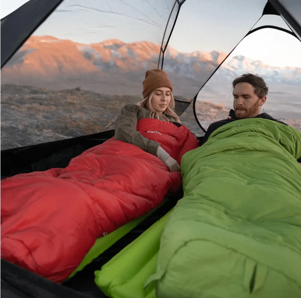 Couple in a tent resting in sleeping bags with a mountain sunrise view, illustrating how to choose a sleeping bag for camping.